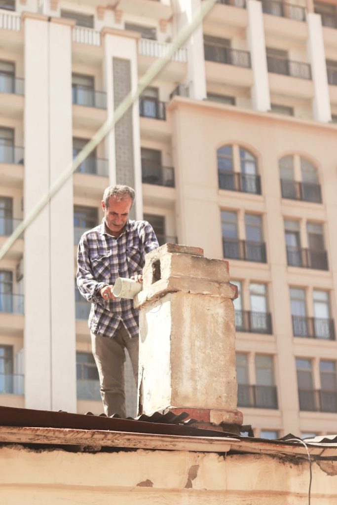 Man in plaid shirt repairing chimney on a city rooftop with modern buildings in background.